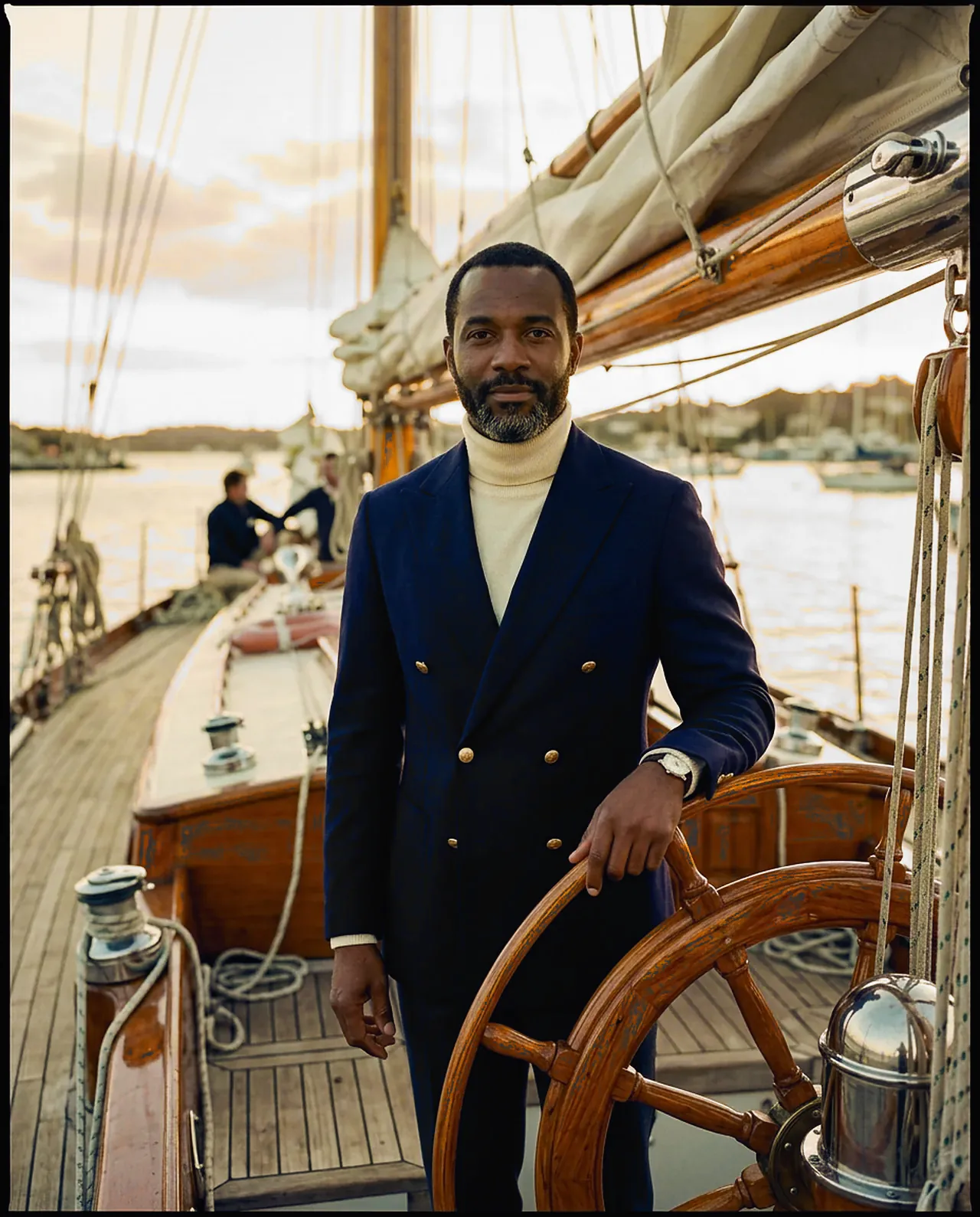 A man in a double-breasted navy blazer and white roll-neck at the wheel of a yacht in harbor.
